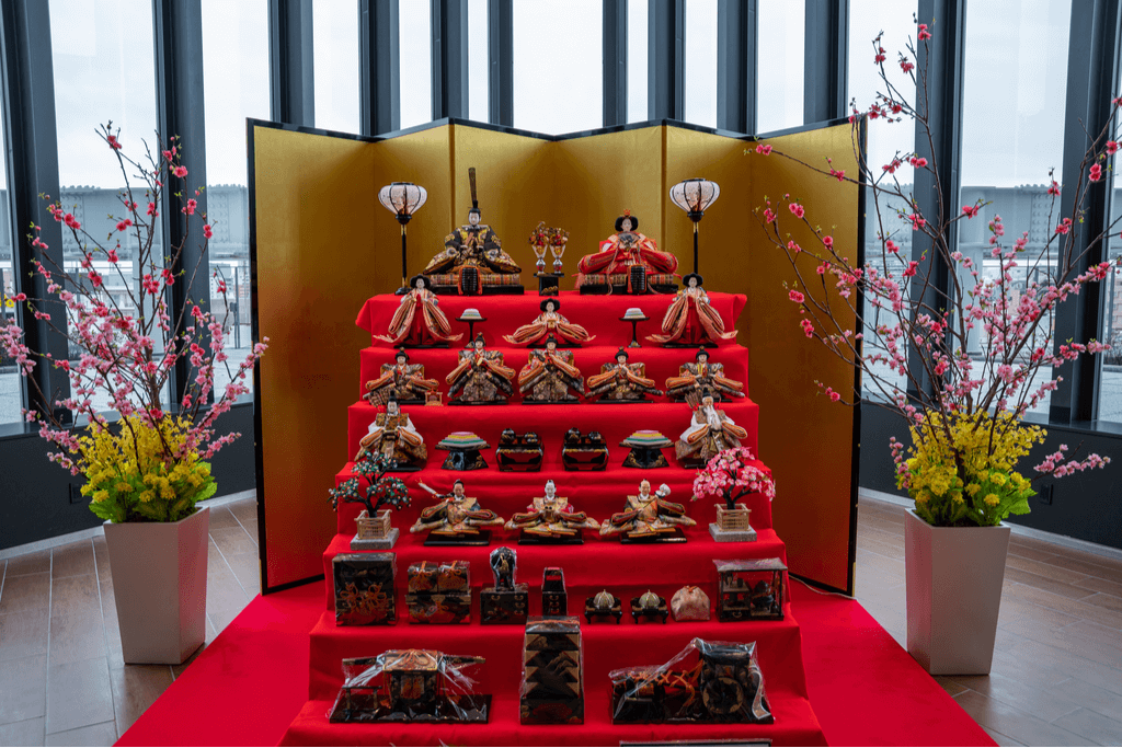 A full display of a Japanese Hina dolls set with all seven traditional tiers in an office building with two sets of flowers on either side in front of many windows.