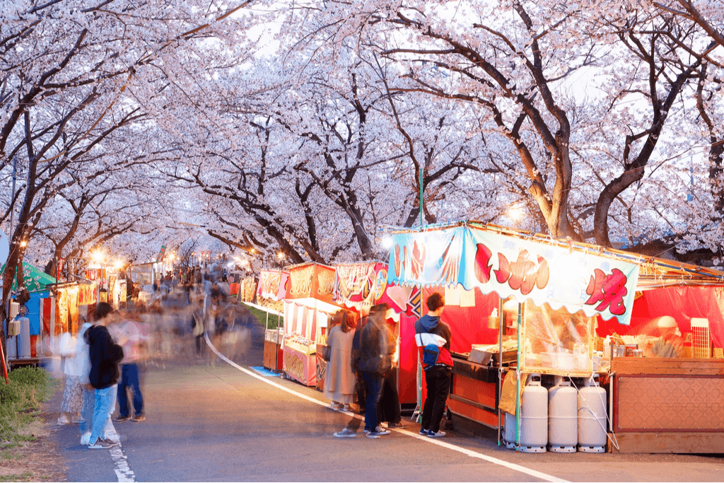 Many food stalls line a walking street with many customers passing through and many cherry blossoms overhead. 