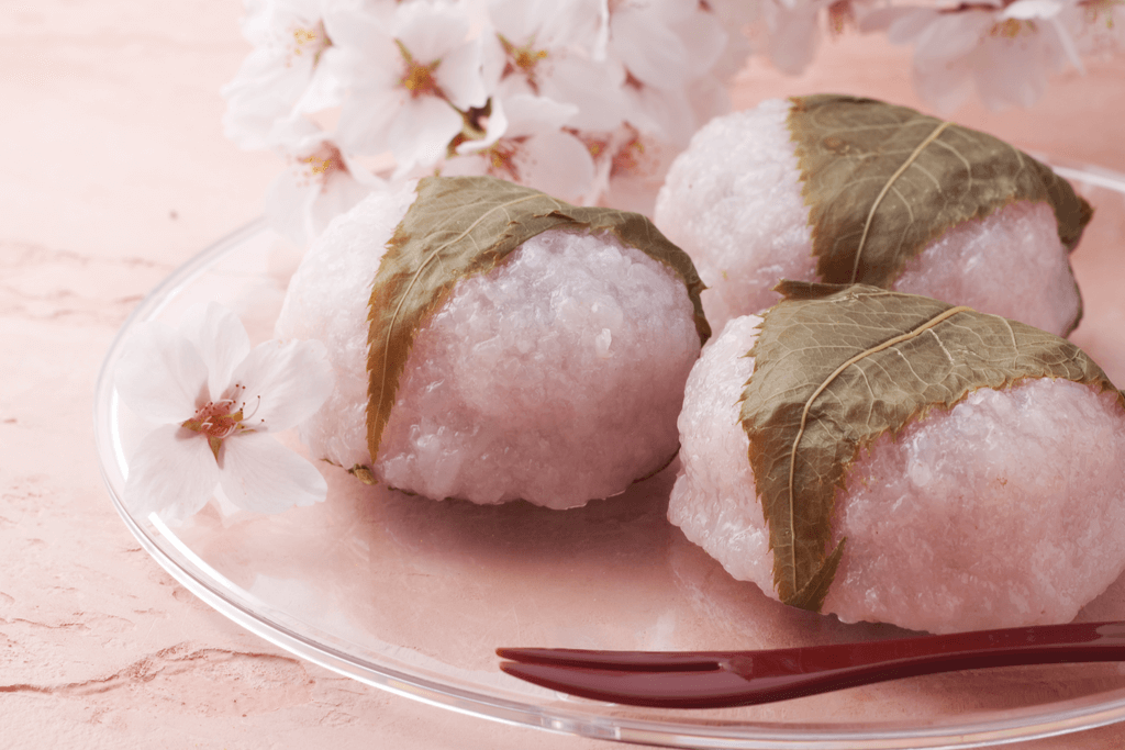 Three servings of sakura mochi, a common sakura food, on a clear plate with cherry blossoms in the background.