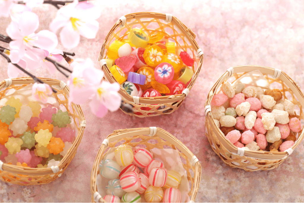 Four baskets, containing Japanese arare and three types of Japanese hard candy, sit on a pink background with cherry blossoms in the foreground.