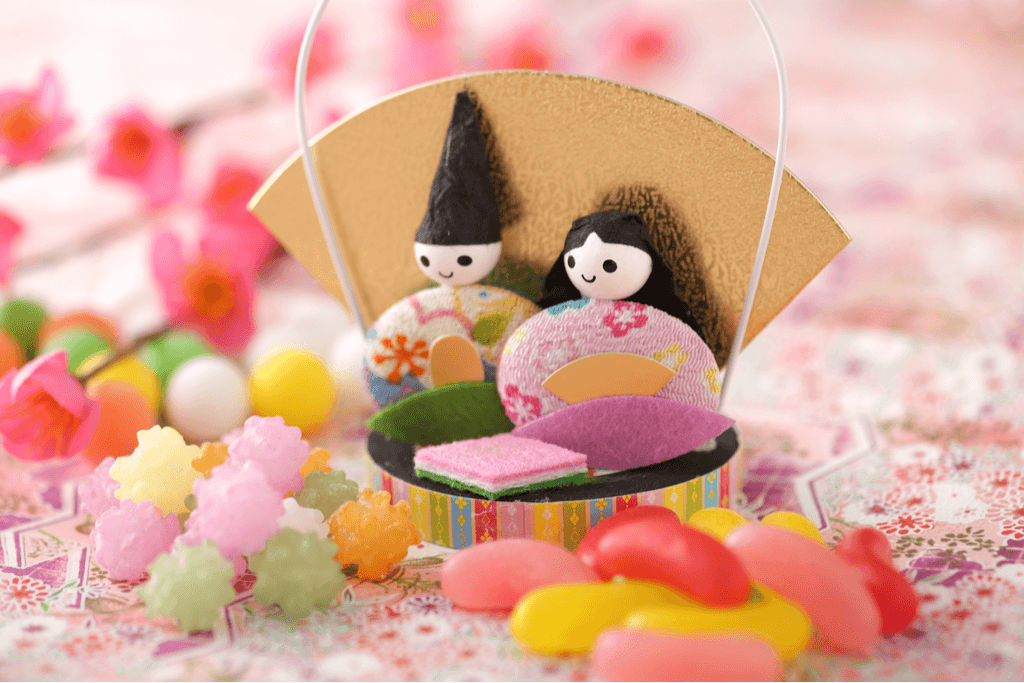 Two simple cloth Hina Dolls sit on a stand surrounded by different hinamatsuri snacks such as jelly beans, konpeito, and other candy.
