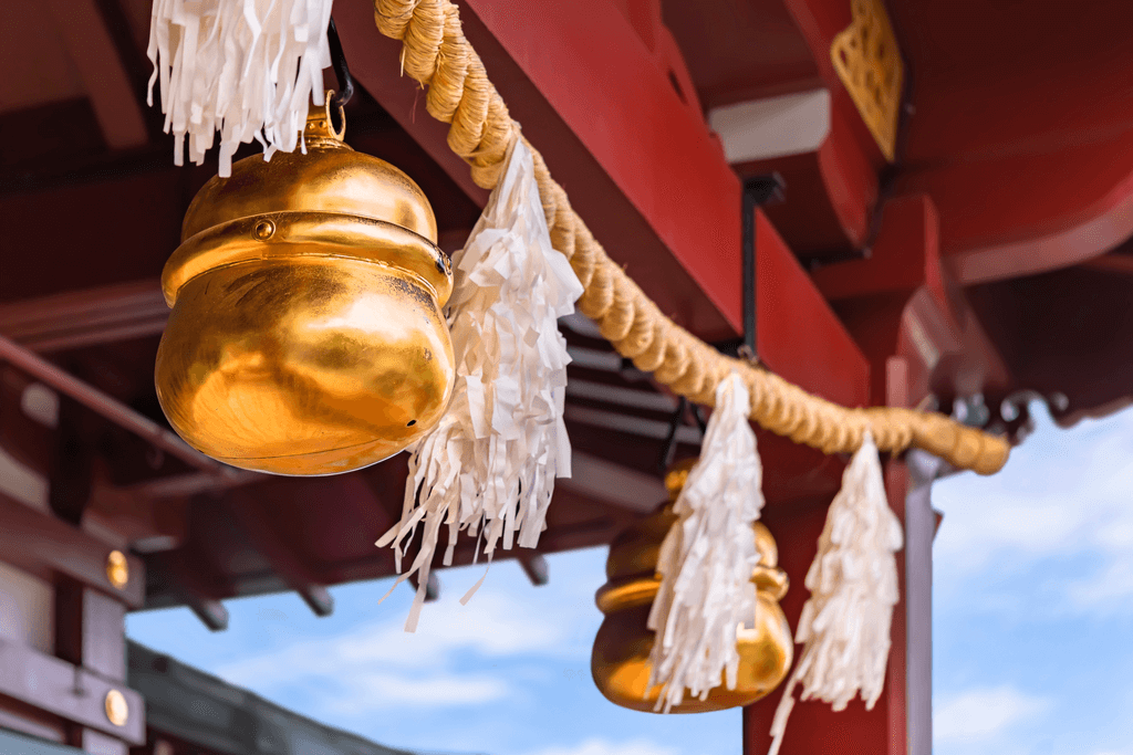 A thick rope, white paper, and golden bells, common symbols at Japanese Shinto shrines, hanging across a part of a shrine.