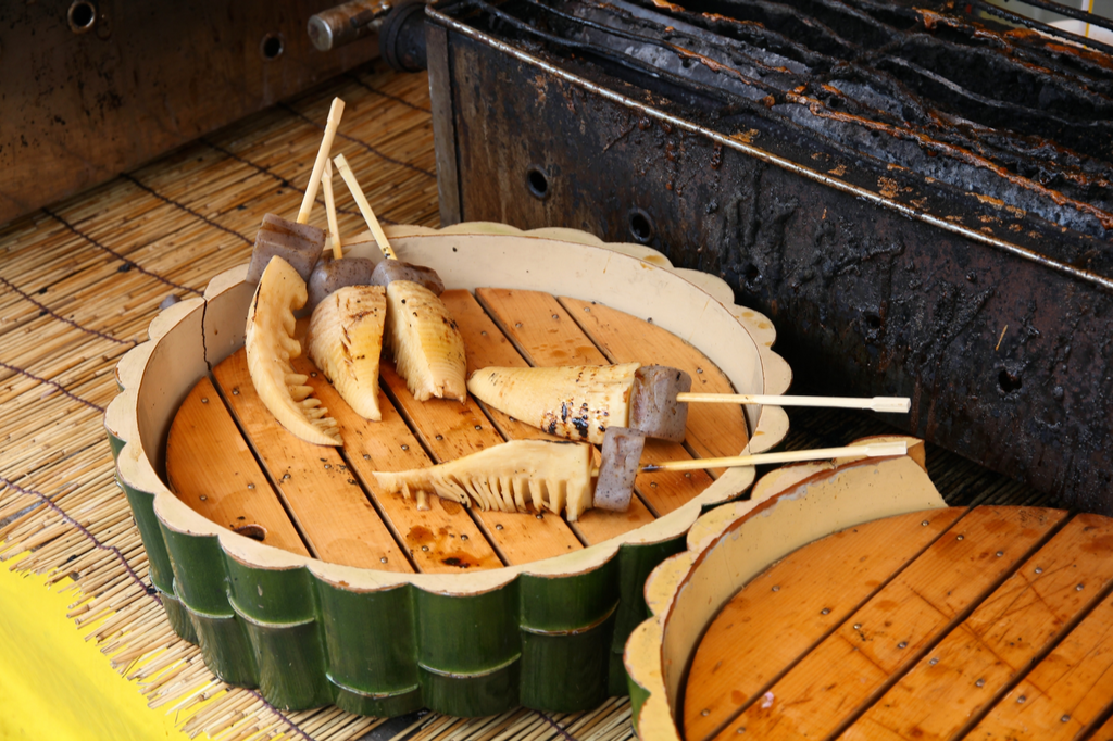 Skewers of grilled bamboo shoots and konjac (a gelatinous vegtable) resting on a circular wooden tub.
