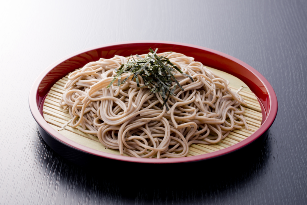 A deep dish plate of zaru soba, which is brown buckwheat noodles on a bamboo strainer, with a small pile of shredded seaweed on top.