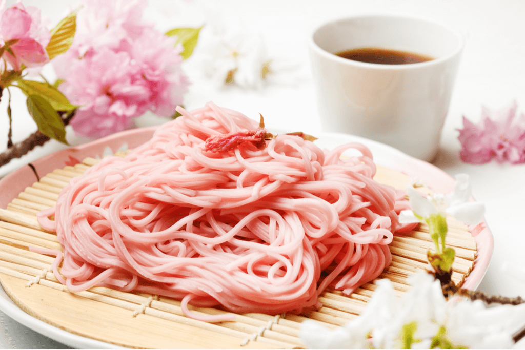 A serving of Japanese sakura cold noodles on a traditional cold noodle serving dish with a bowl of the dipping sauce or soup on the side and sakura petals all around.