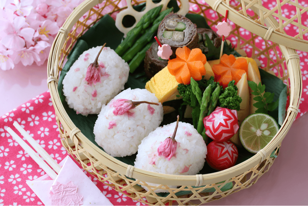 A basket sits on a table filled with different vegetables cut into sakura shapes and small rice balls made with sakura petals, a popular sakura food.