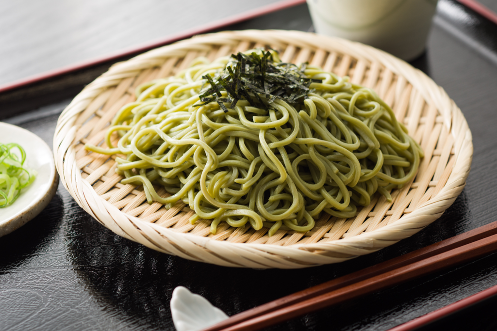 A pile of green tea noodles on a soft bamboo strainer, with a pair of brown chopsticks on the side.