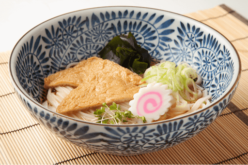 A blue and white porcelain bowl of kitsune soba, which includes noodles, scallions, fish cake, kombu seaweed and of course, triangle-shaped fried tofu.