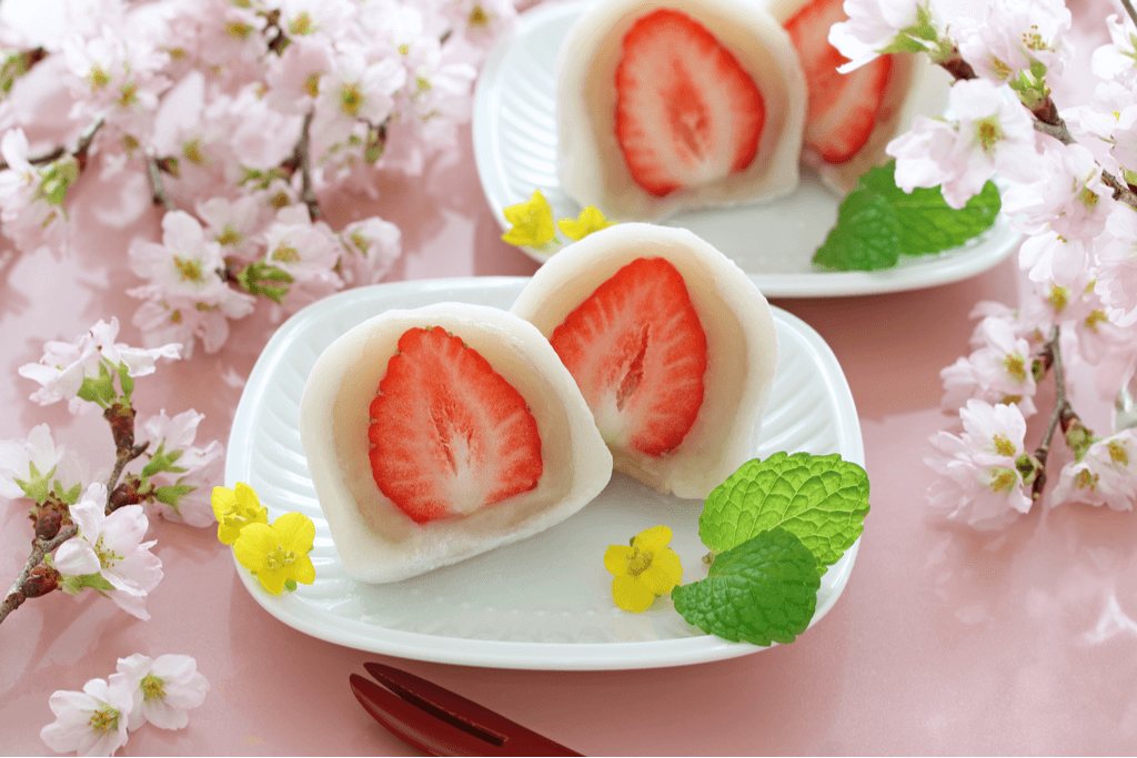 Two plates of strawberry daifuku, one of the types of daifuku that uses a white bean paste, cut in half showing the layers of strawberry, white bean paste, and mochi.