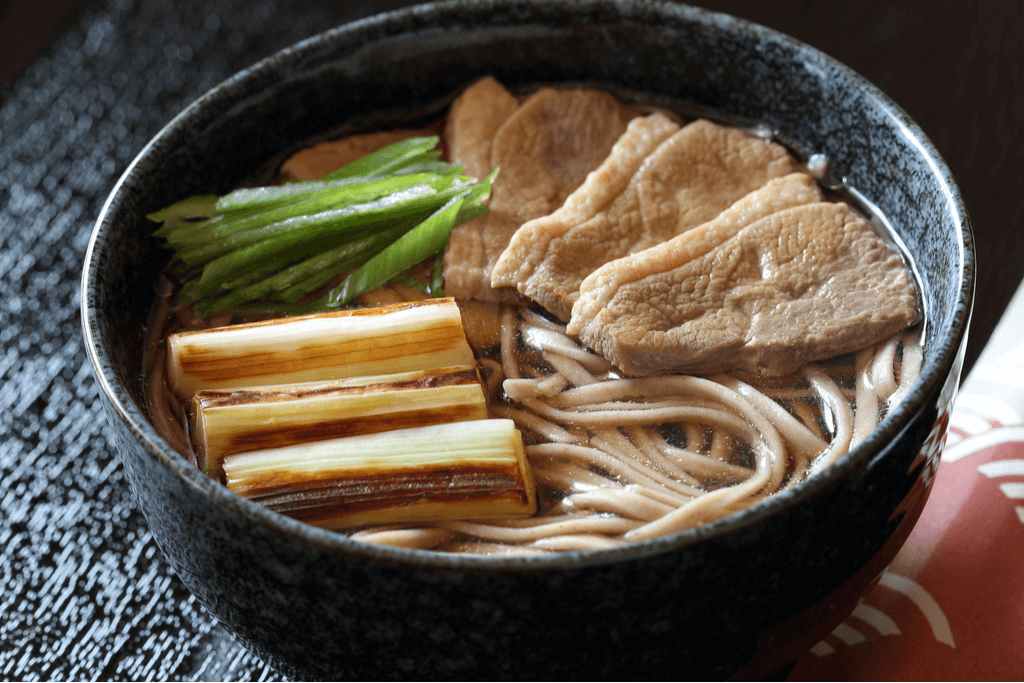 A bowl of Nanban soba, one of the types of soba featuring a duck and green onion topping, in a black bowl on a black table.