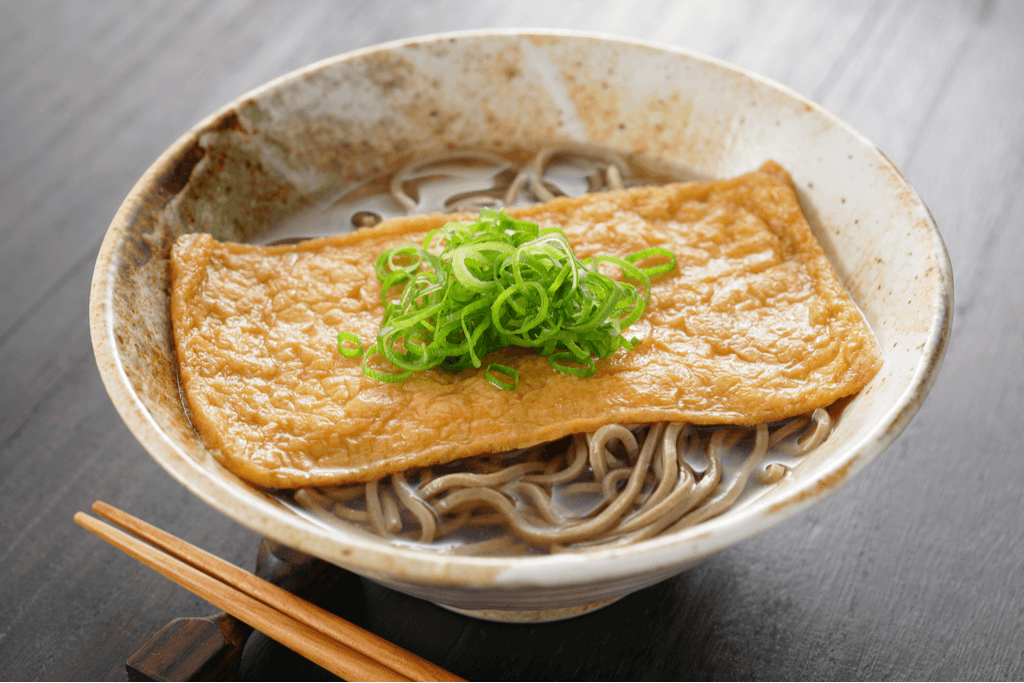 A bowl of kitsune soba, one of many types of soba dishes, which features a piece of fried tofu on top of hot soba with green onion slices on top.