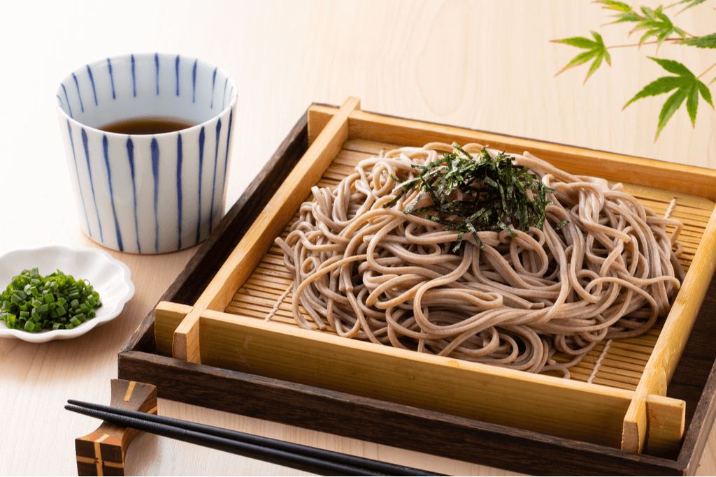 A serving of zaru soba, one of the most basic types of soba dishes, on a bamboo tray with seaweed on top and green onion and dipping sauce on the side.