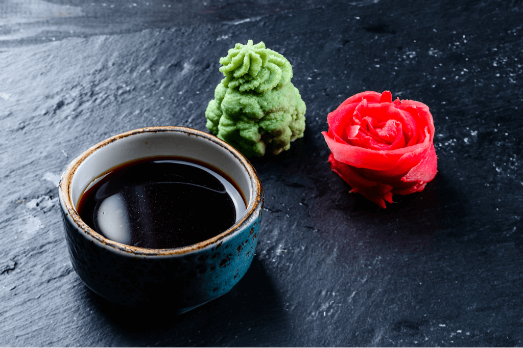 A small dipping bowl of soy sauce next to an aesthetic pile of wasabi and red ginger shaped like a flower on a black stone surface.