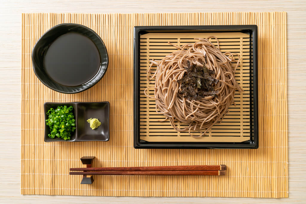 A placemat with zaru-style soba noodles on it, with the noodles topped with seaweed strips on a zaru tray, a holder of chives and wasabi, and a bowl of dipping soup.
