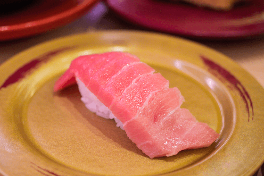 A piece of tuna sushi sits on a yellow plate at a sushi restaurant with a pinker hue.