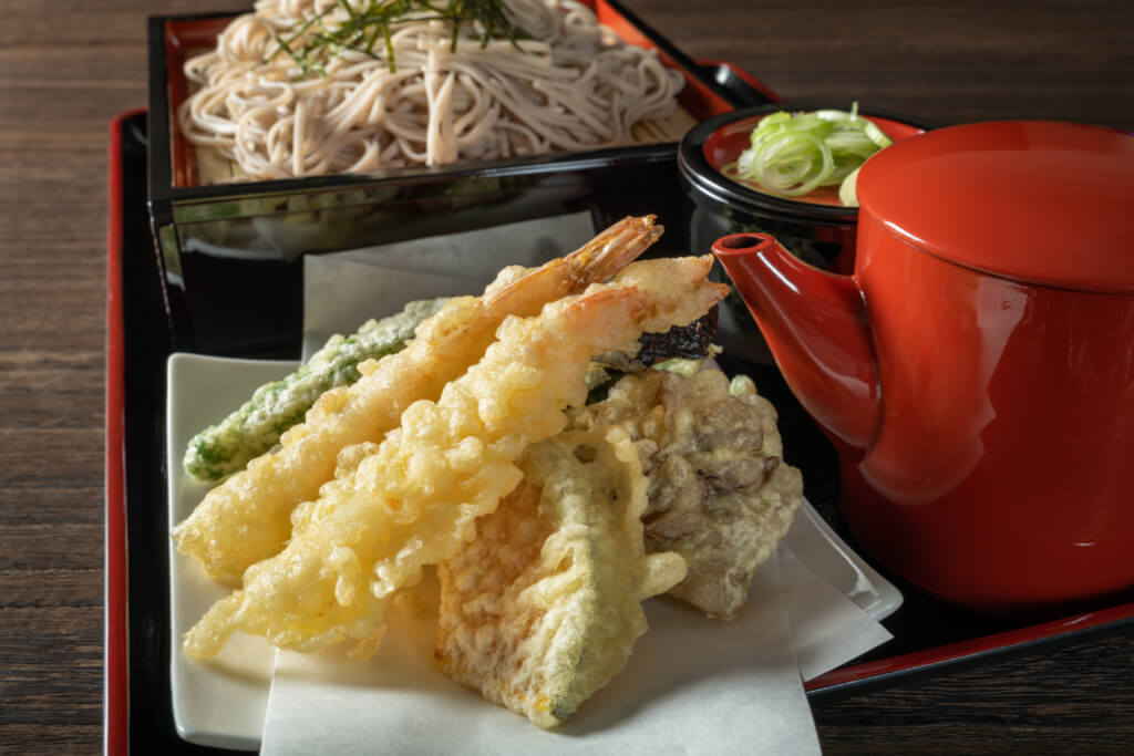 Tenzaru soba, one of the most popular cold soba dishes out there, on a tray with delicious shrimp and vegetable tempura next to soba noodles, and a red container of tempura sauce with a spout.