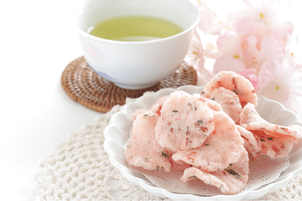 A plate of cherry blossom senbei, sakura-flavored snacks made of shrimp and other ingredients, on a white plate with a cup of tea and cherry blossoms in the background.