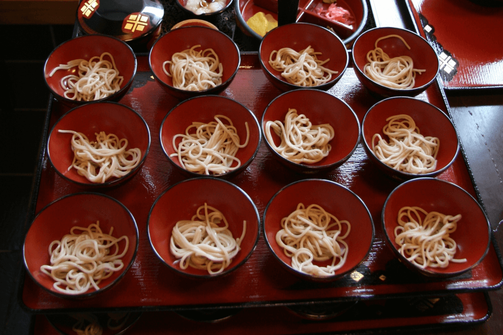 Many bowls of wanko soba, one of the types of soba named after its small bowl, on a tray with toppings of pickled vegetables in the background.