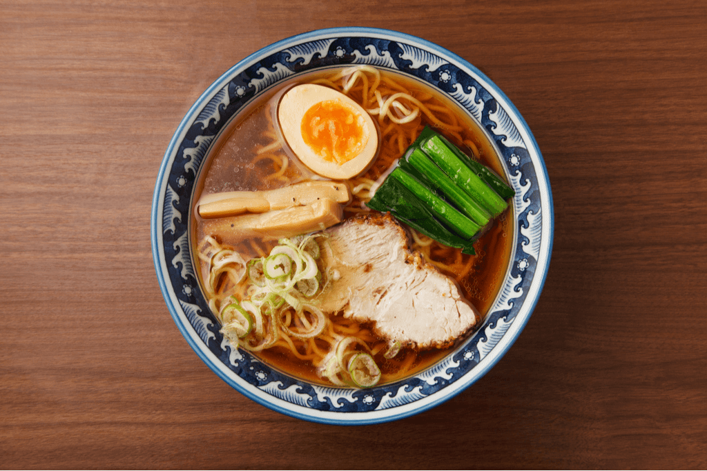 A blue bowl filled with shoyu ramen with a boiled egg, water chestnut, green onion, spinach, and meat as a topping on a wooden background.