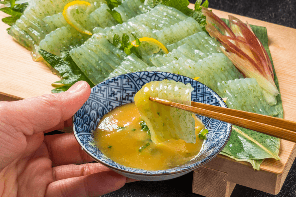 A wooden board with kelp, onion, and konjac jelly sashimi with a grooved texture on top, and a pair of hands dipping the konjac sashimi into a yellow yuzu sauce in a blue dipping saucer.