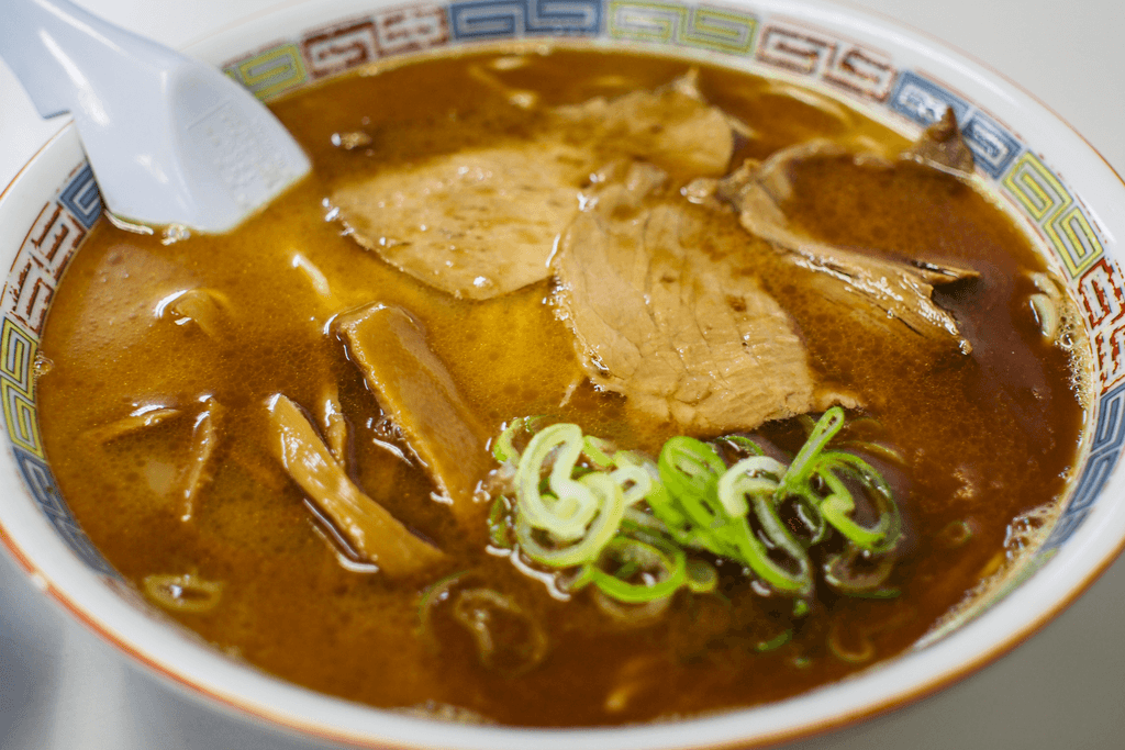A bowl of shoyu ramen using a beef stock for the broth and is a darker brown, topped with water chestnuts, pork belly, and green onions.