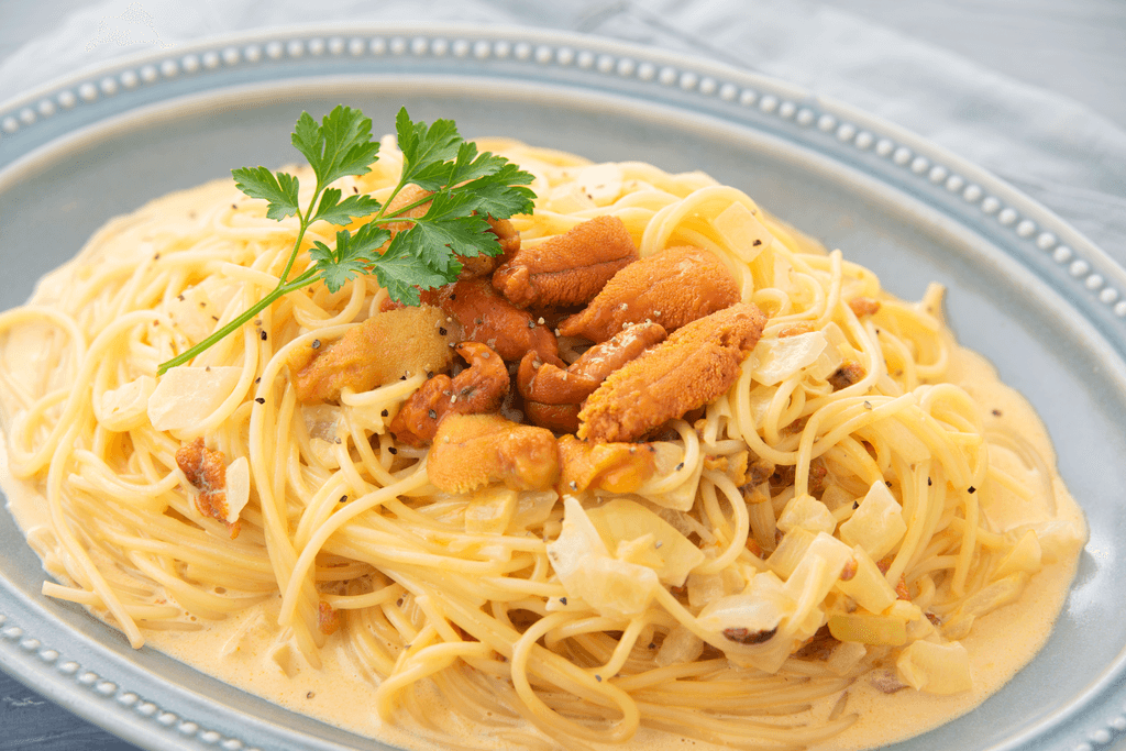 A plate of pasta sits on a table, with the pasta being spaghetti in a cream sauce with orange, cooked bits of uni, onion, and herbs on top.