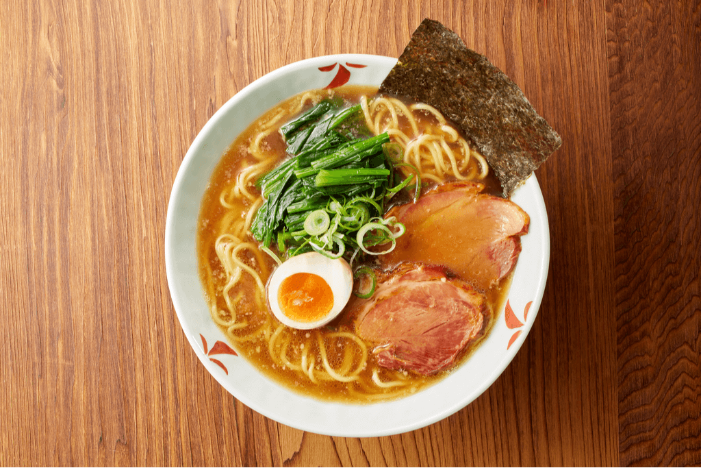A white bowl on a wooden table holds a bowl of ramen noodles that combines pork bone and shoyu ramen with pork belly, egg, seaweed, green onion, and seaweed as toppings.