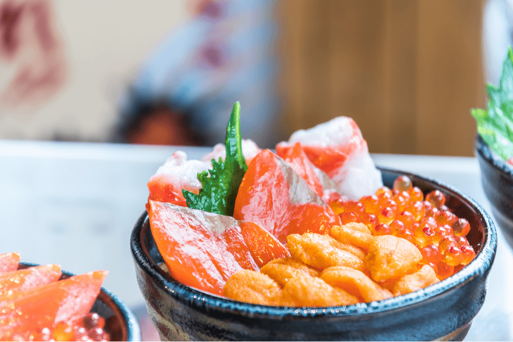 A colorful Japanese sashimi rice bowl sits on a table next to two others on the side, with colorful crab, salmon, tuna, salmon roe, and uni on top of the rice. 