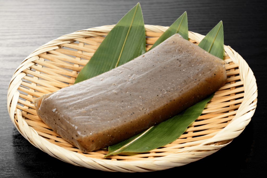 A block of green-gray konjac jelly sits on a bamboo tray and kelp leaves on a black table.