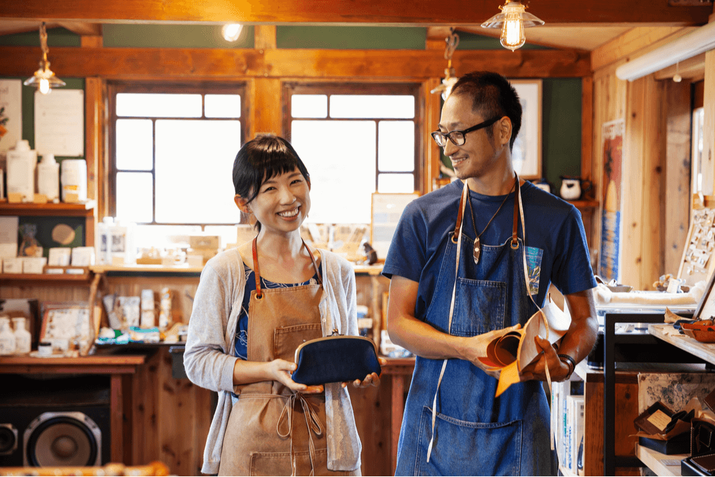 A Japanese man and woman wearing aprons, smiling and holding shoes they just made. They are local Japanese makers.