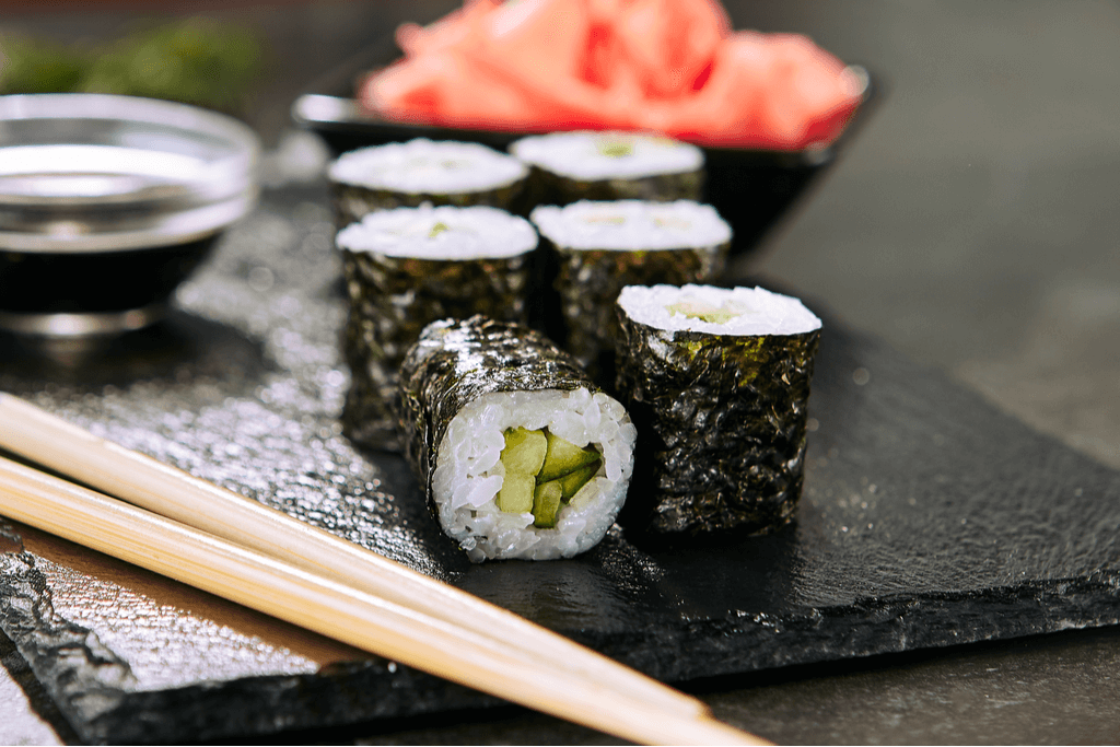 Rolls of Japanese kappa sushi on lay on a black serving slab with pickles, soy sauce, and chopsticks around it on a black table.