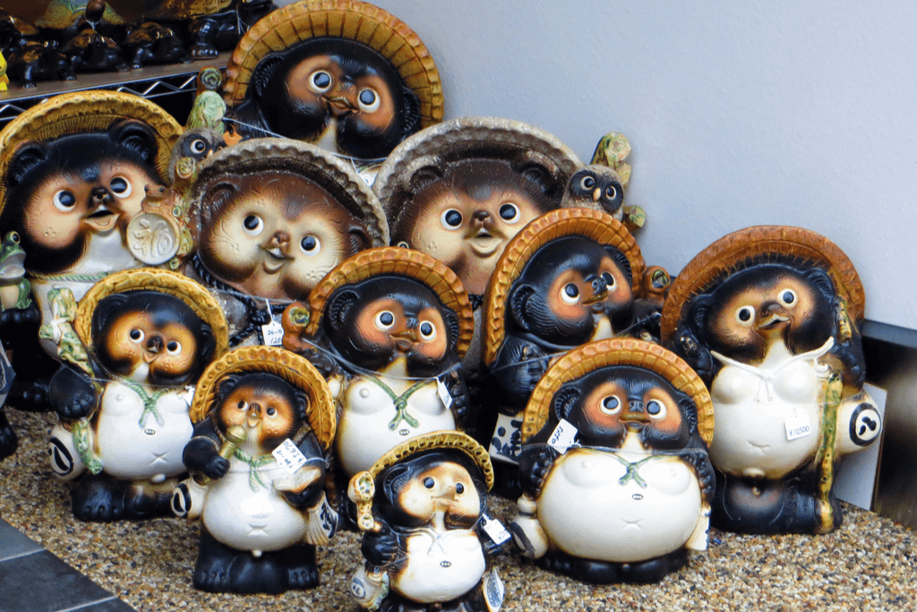 Eleven tanuki statues stand in a shop with tags on each one in varying sizes, poses, and expressions, all with hats, sake bottles, and other normal tanuki symbls.