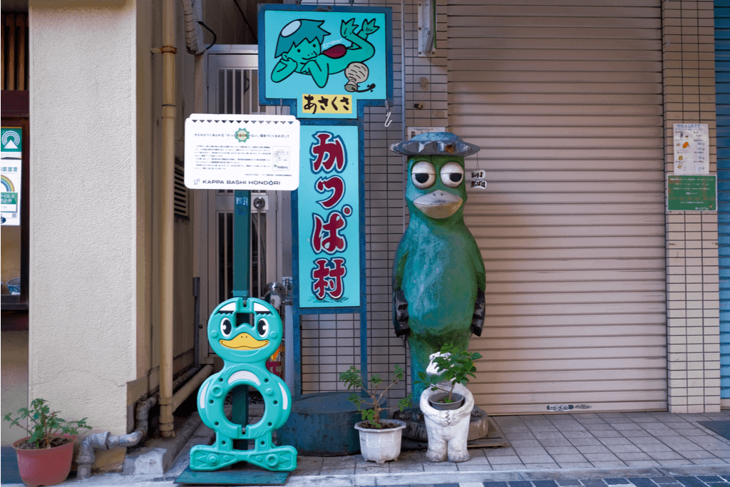 Three Japanese kappa items: a statue, a sign with a cute illustration and text for Asakusa's Kappa Village, and a barrier end, stand in front of a building with a closed gate.  