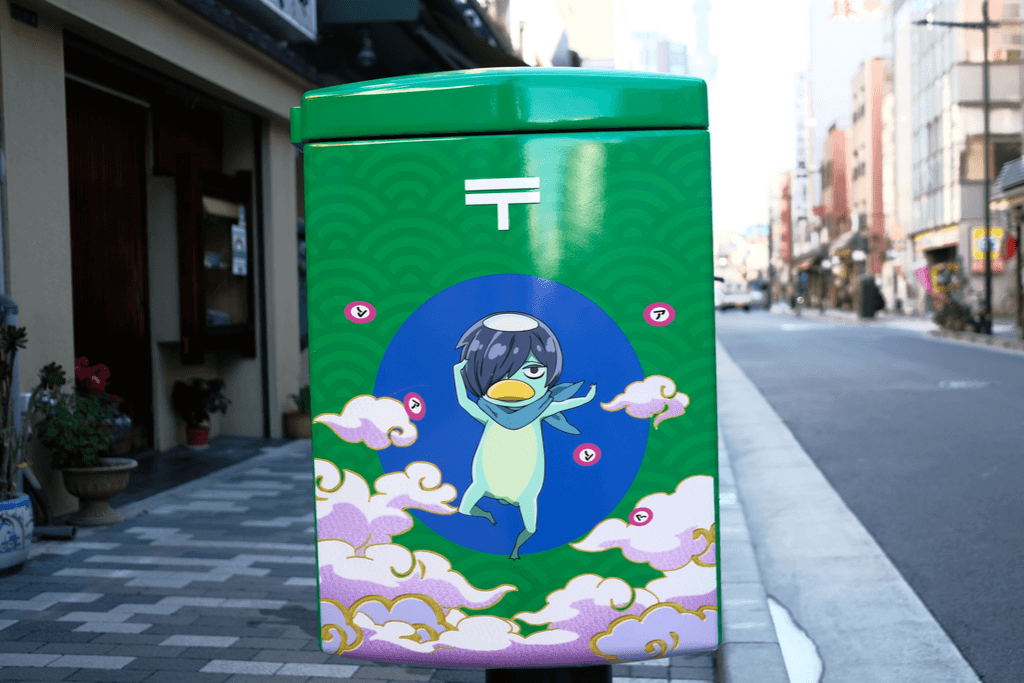 A cute illustration of a Japanese kappa with a bandana in front of a blue circle and steam decorates a Japanese post box on a street in Japan.