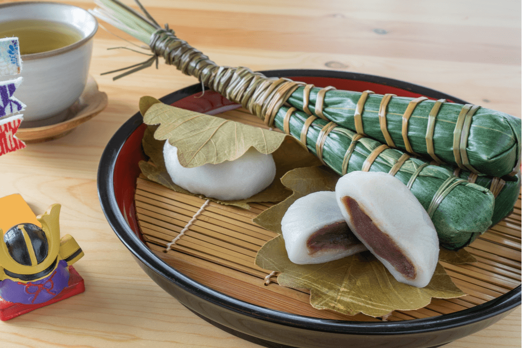 Four pieces of Children's Day snacks, two pieces of Kashiwamochi, which is a rice cake stuffed with red bean paste and placed in a leaf, and Chimaki, a piece of mochi wrapped in a leaf and tied with dried leaf, on a Japanese bamboo tray next to a ceramic samurai hat.