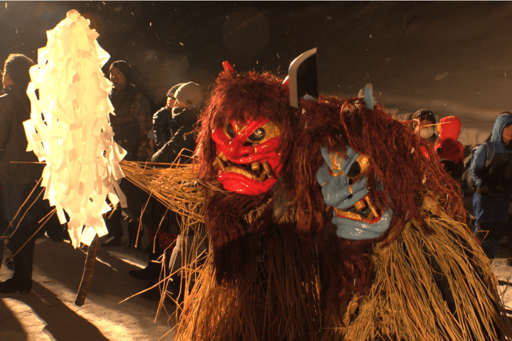 Two people dressed as oni, or Japanese ogres, with red and blue oni masks, straw clothes, red hair, and a fake staff made of paper stand outside with festival-goers in the background. 