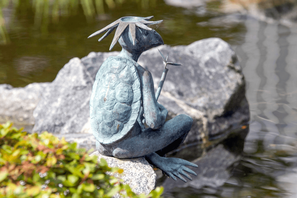 A bronze statue of a Japanese kappa, one of the more malicious Japanese mythical creatures, sits on a rock next to a body of water as it holds a reed.