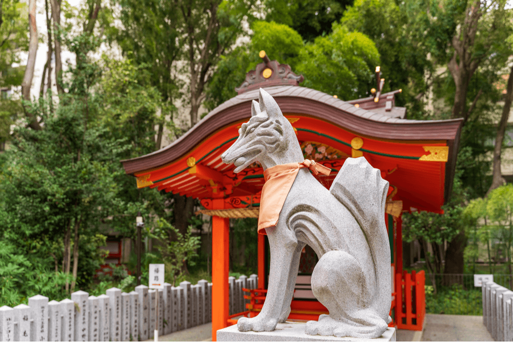A statue of a fox, a powerful Japanese mythical creature, sits in front of a small shrine with many trees around it.