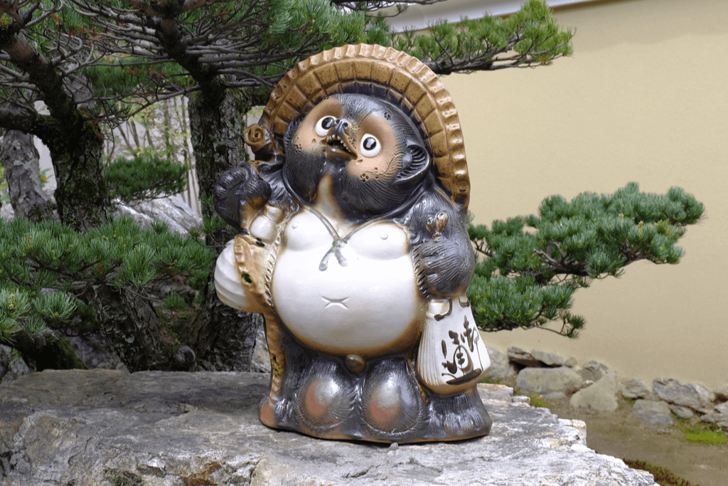 A statue of a tanuki, or Japanese raccoon dog,  holds bag and sake while wearing a hat, stands on top of rock in the garden area of a Japanese restaurant.