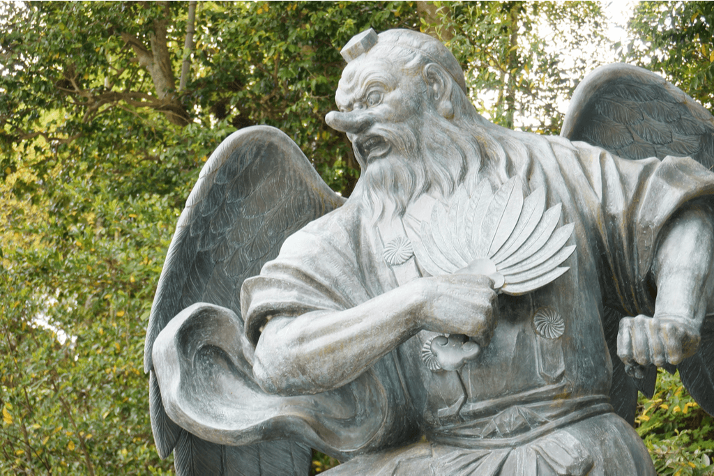 A statue of a Tengu, Japanese mythical creatures that look like bird men, stands in an aggressive pose with its wings out and a feather fan in his hand with an angry look on his face.