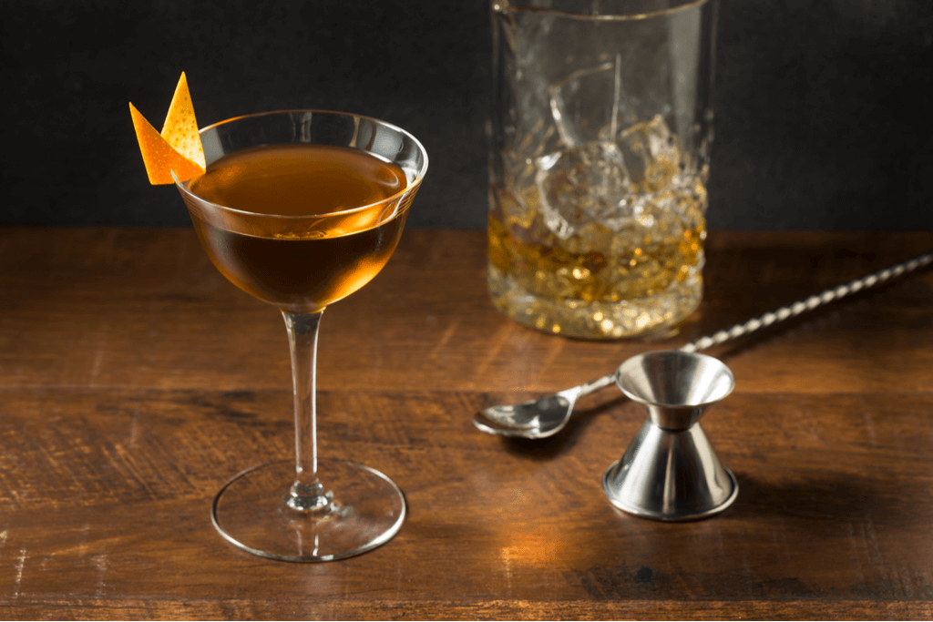 A cocktail glass holds Bamboo, a Yokohama cocktail, with an orange peel used as garnish on the glass, next to another glass, a stirrer and a measuring cup.