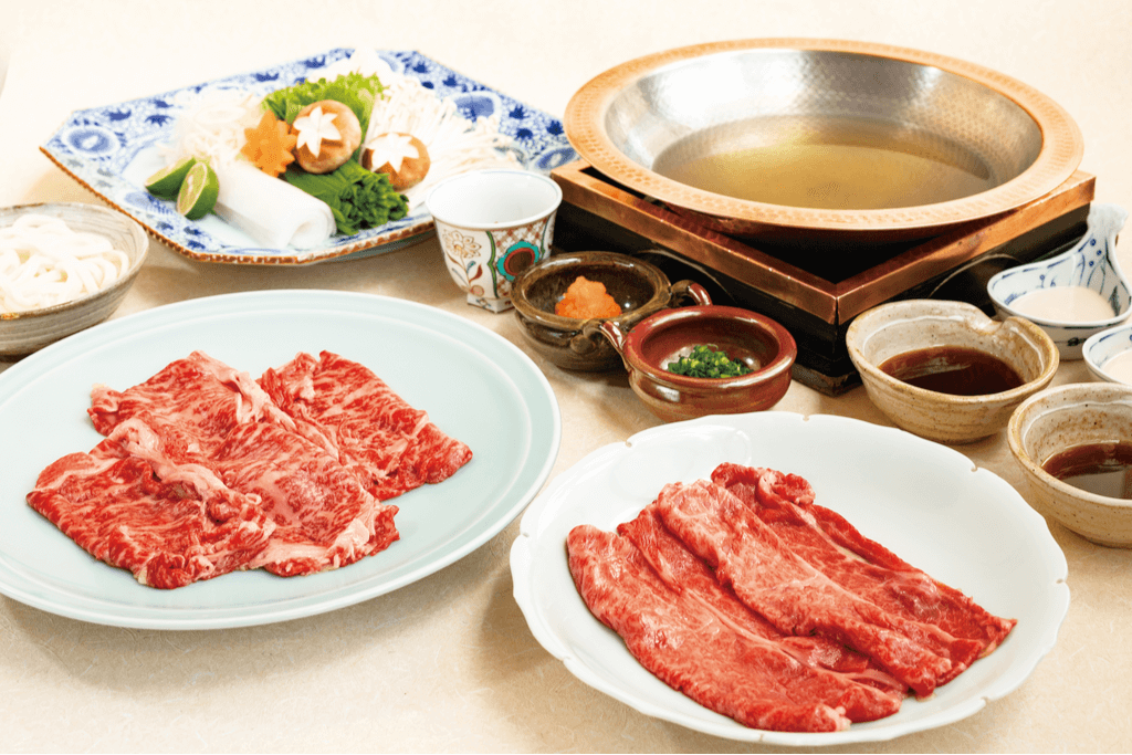 A table with a spread of Gyunabe, one of the oldest Yokohama foods, with plates of sliced beef, vegetables, noodles, sauces, green onions, and a hotpot with a soup.