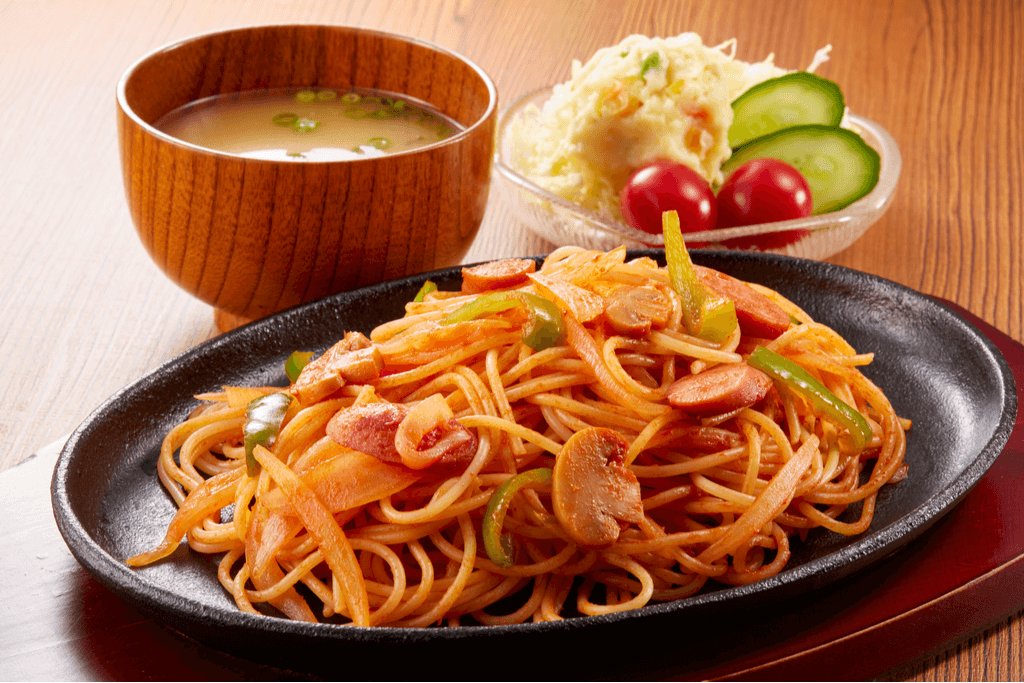 A black plate of Napolitan, one of the Western-inspired Yokohama foods, with spaghetti, onions, sausage, mushrooms, and peppers in a red sauce, next to a bowl of miso soup and potato salad.