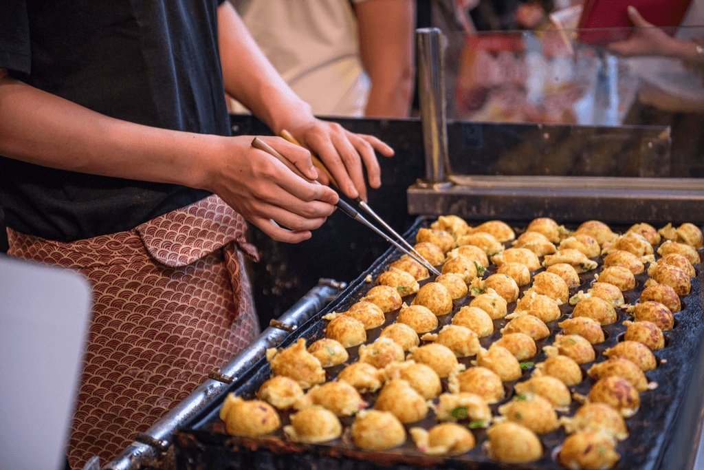 One of the Japanese street vendors at a street market uses chopsticks to flip the grilled octopus balls on his cooking station. 
