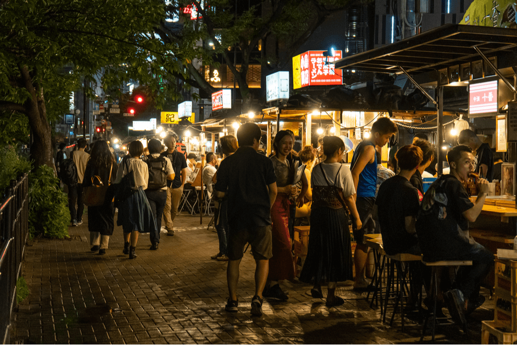 Many young people walk down the street past Japanese street vendors stalls, where people are eating a variety of foods on a summer night.