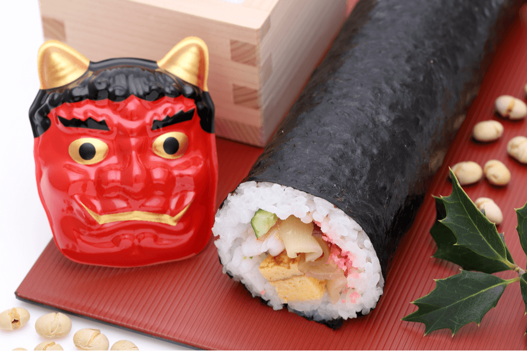 A Japanese oni mask, which is red and has black eyebrows and hair and gold eyes, tusks, and horns, next to a lucky sushi roll and beans on a red tray.