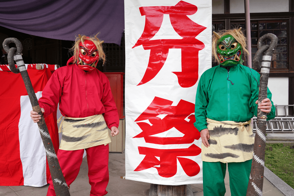Two men dressed as Japanese oni, one in red and the other in green, hold two giant oni clubs while standing in front of a sign for a Setsubun festival.