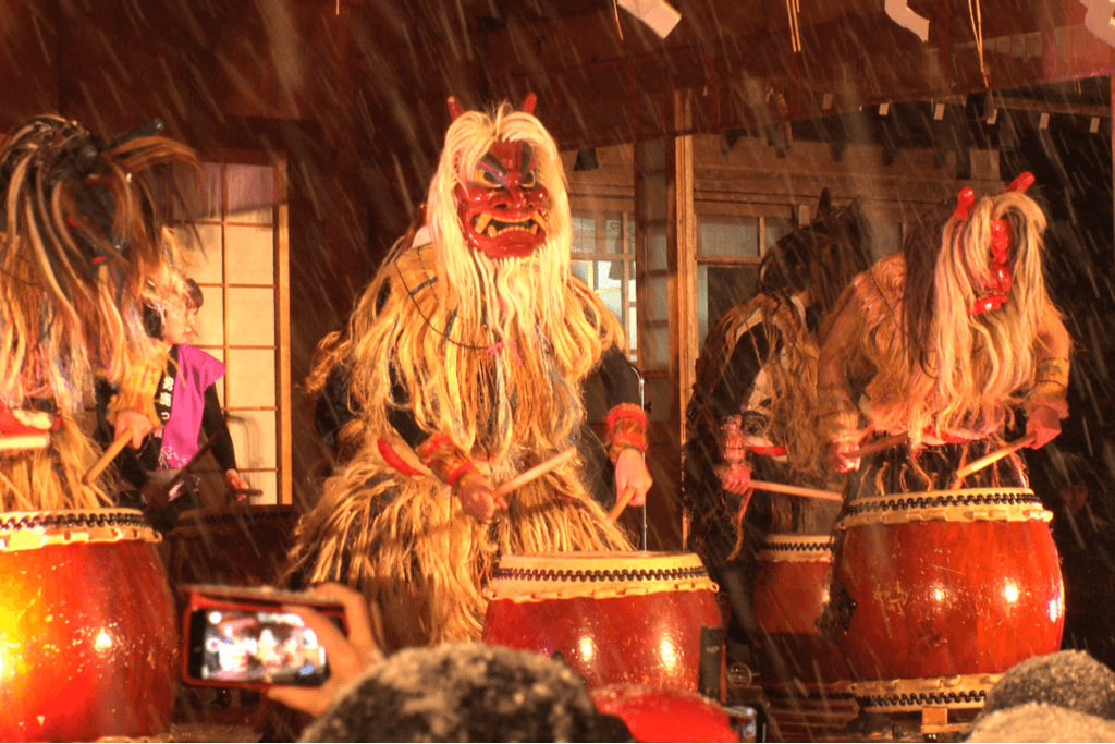 Three men in Japanese oni masks and a traditional garb made of animal hair as they play the Japanese drums on a stage at a Japanese-style building.