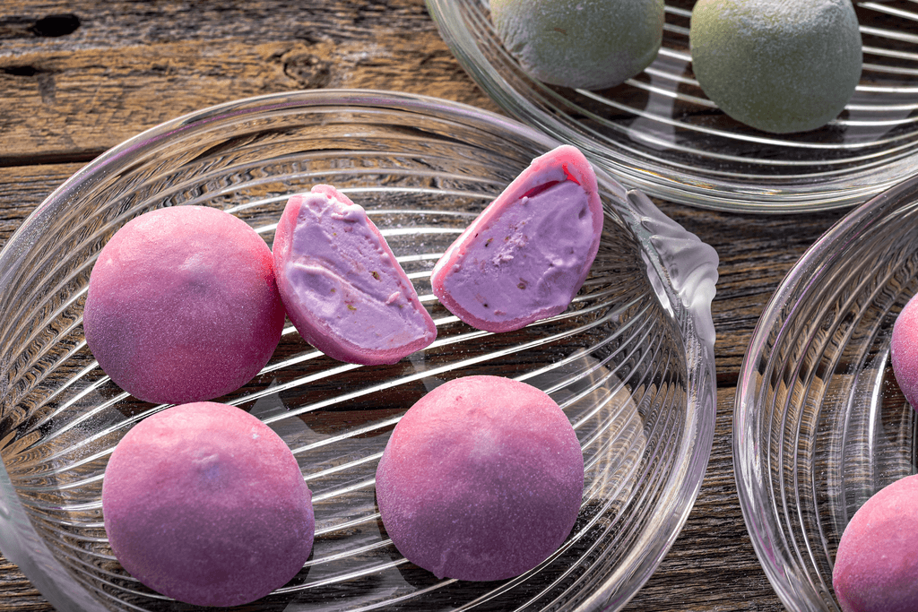 Plates of mochi ice cream sits on a wooden table with one of the types of mochi ice cream being cut in half showing purple mochi and ice cream in a strawberry flavor