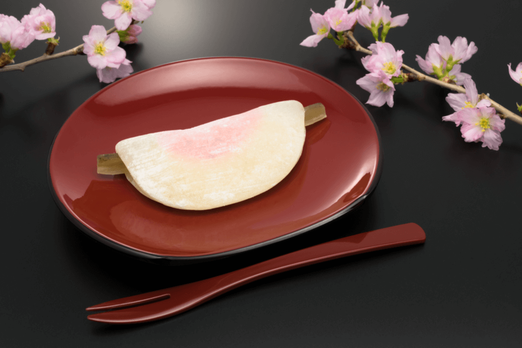 A red plate holds Hanabira mochi, one of the types of mochi eaten around New Years, with a piece of bamboo hanging out of either sides of the mochi and a red utensil and plum blossoms next to it on a black table